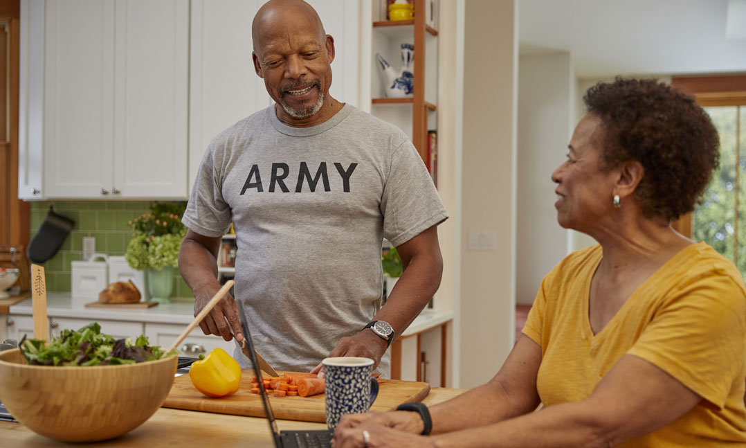 Army veteran chops vegetables at counter while talking with wife who is on the laptop 